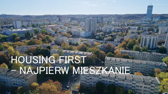 Plansza tytułowa filmu "Housing First - najpierw mieszkanie". Blokowisko filmowane z lotu ptaka.