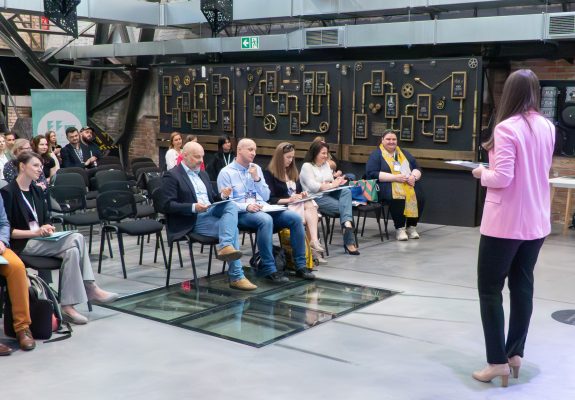 Zdjęcie przedstawia widownię Demo Day śląskiej przestrzeni innowacji i prelegentkę z zaangażowaniem opowiadającą o czymś publiczności.