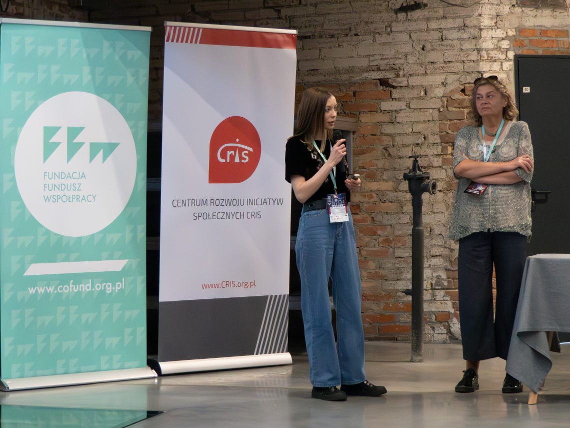 Prelegentka wydarzenia mówi do mikrofonu na tle logotypów partnerów projektu.