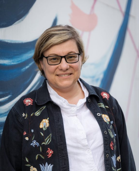 portret na tle muralu uśmiechniętej od profilu kobiety w koszuli i narzuconej czarnej kurtce wyszywanej kwiatami