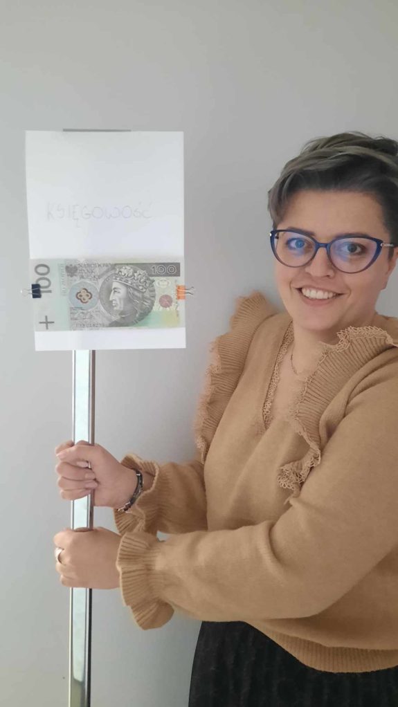 uśmoiechnięta kobieta o krótkich włosach w żółtej koszuli trzyma baner składający się z rurki pcv i przyklejonej do niej kartki a4 z napisem "księgowosć" i przyczepionym na klipsy banknotem 100złotowym