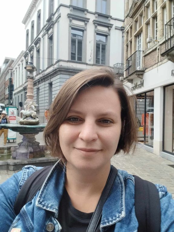Kobieta z grzywką w dżinsowej kurtce na tle secesyjnych budynków na ulicy