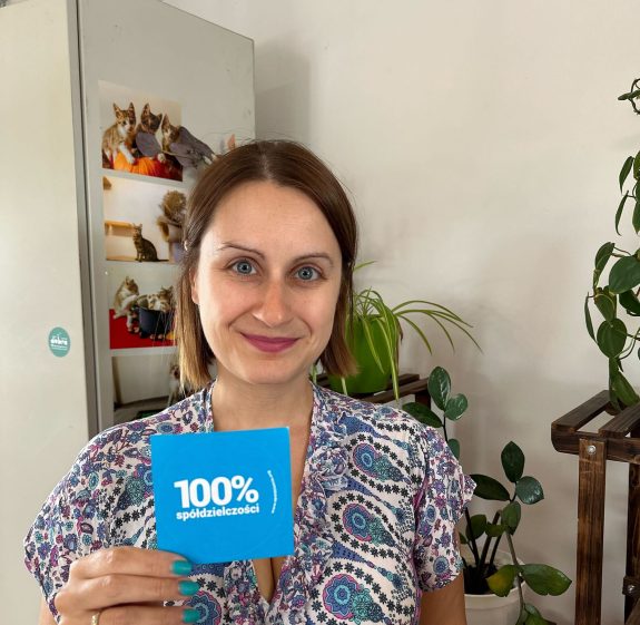 usmiechnięta kobieta trzymająca małą kartkę tego samego koloru, co oczy z napisem 100% spółdzielczości