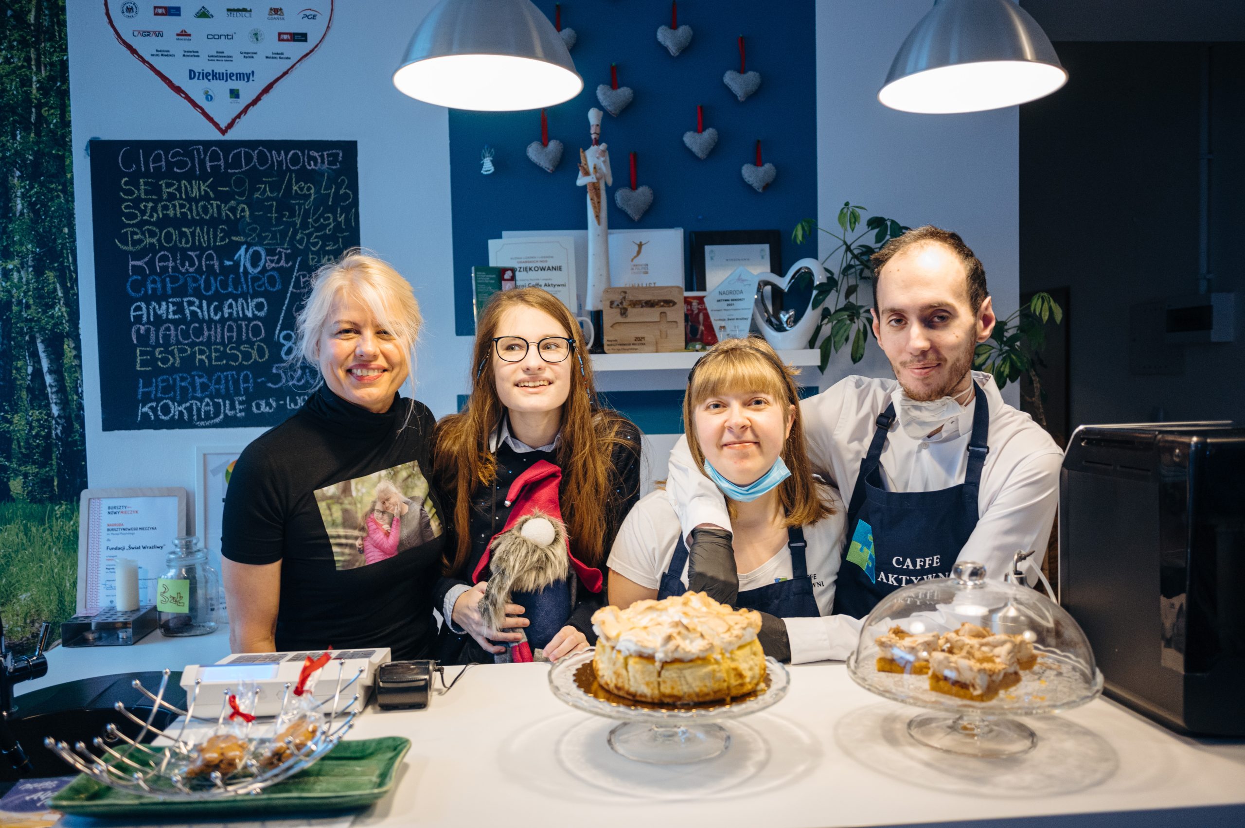 czworo baristów w fartuchach opartych o biala lade, na ktorej stoja dwa ciasta